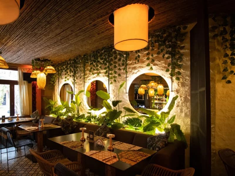A warmly lit dining room: timber tables under glowing pendant lamps, ivy trailing across textured plaster walls