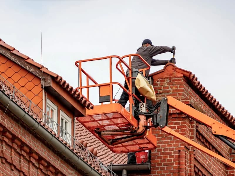A roofer working from a lift platform against the brick gables and tiled roof of a house