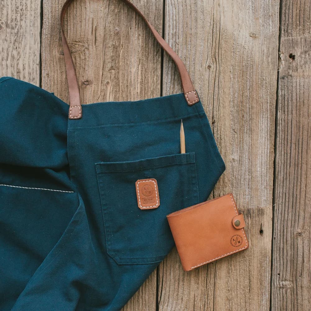 A folded waxed-canvas apron with leather ties, hanging against a plain wall