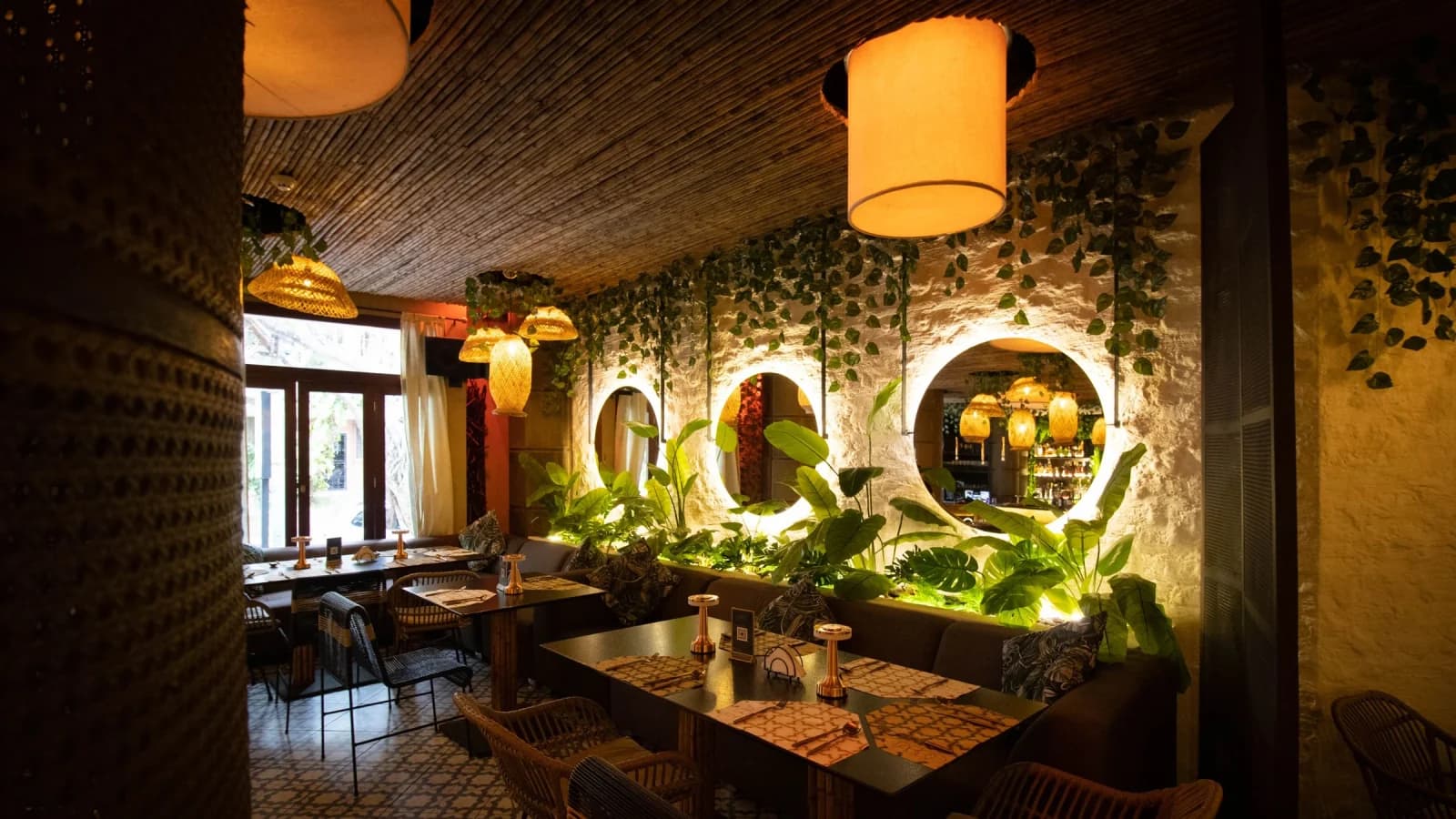 A warmly lit dining room: timber tables under glowing pendant lamps, ivy trailing across textured plaster walls